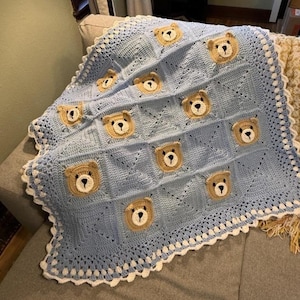 May include: A handmade baby blanket in light blue, featuring a repeating pattern of crocheted teddy bear faces. The blanket has a white scalloped border and is made of soft yarn. A cozy and comforting item for a nursery.