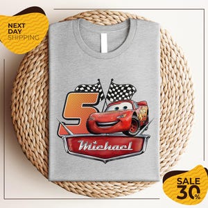 A gray t-shirt with a graphic design featuring a red car and checkered flags, along with the text "Michael" and the number "5". The t-shirt is displayed on a woven placemat, and there are yellow price tags indicating a sale of 30% off.