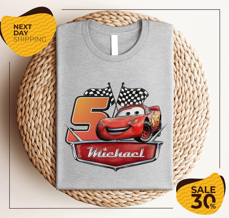A gray t-shirt with a graphic design featuring a red car and checkered flags, along with the text "Michael" and the number "5". The t-shirt is displayed on a woven placemat, and there are yellow price tags indicating a sale of 30% off.