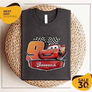 A gray t-shirt with a graphic design featuring a red car and checkered flags, along with the text "James" and the number "9". The t-shirt is displayed on a woven placemat, and there are yellow price tags indicating a sale of 30% off.