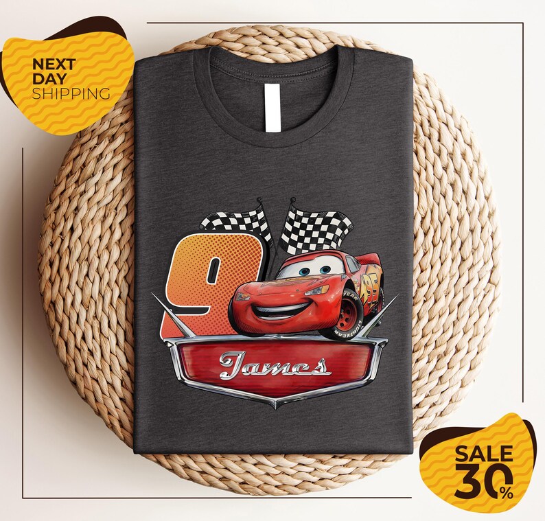 A gray t-shirt with a graphic design featuring a red car and checkered flags, along with the text "James" and the number "9". The t-shirt is displayed on a woven placemat, and there are yellow price tags indicating a sale of 30% off.