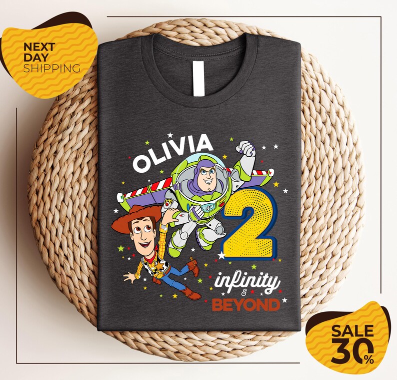 a gray t-shirt with a design of two characters from the Toy Story franchise, specifically Buzz Lightyear and Woody, along with the text "Olivia 2 infinity & beyond". The t-shirt is displayed on a woven placemat, and there are two yellow price tags in the image, one on the left and one on the right, indicating a sale or discount.