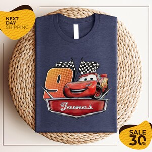 A blue t-shirt with a graphic of a red car and checkered flags, along with the text "James" and the number "9". The t-shirt is displayed on a woven placemat, and there are yellow price tags with text indicating a sale.