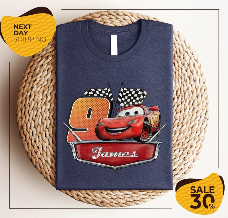 A blue t-shirt with a graphic of a red car and checkered flags, along with the text "James" and the number "9". The t-shirt is displayed on a woven placemat, and there are yellow price tags with text indicating a sale.
