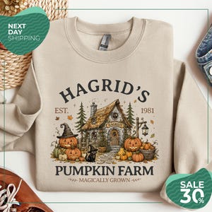 May include: A beige crewneck sweatshirt featuring a graphic print of a stone cottage with a thatched roof, surrounded by pumpkins, pine trees, and a black cat wearing a witch hat. The text reads HAGRID'S EST. 1981 PUMPKIN FARM MAGICALLY GROWN.