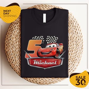 A black t-shirt with a graphic design featuring a red car and checkered flags, along with the text "Michael" and the number "5". The t-shirt is displayed on a woven placemat, and there are yellow price tags indicating a sale of 30% off.