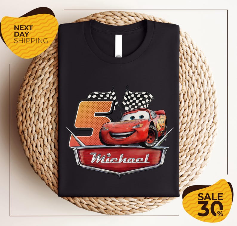 A black t-shirt with a graphic design featuring a red car and checkered flags, along with the text "Michael" and the number "5". The t-shirt is displayed on a woven placemat, and there are yellow price tags indicating a sale of 30% off.