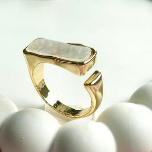 Könnte beinhalten: Ein goldfarbener offener Ring mit einer rechteckigen, schimmernden weißen Einlage auf der Oberseite. Die Ringschiene hat eine glatte, polierte Oberfläche mit einer Lücke zwischen den Enden und zeigt eine klare Metallstruktur mit flacher Fassung.