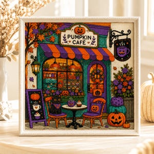 May include: A framed cross-stitch embroidery depicting a Pumpkin Café storefront with a purple and orange striped awning, a black cat sitting on a chair, jack-o'-lanterns, a menu board, and floral decorations in shades of orange, purple, and green.