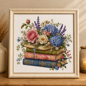 May include: A framed cross-stitch embroidery featuring a stack of four vintage-style books in cream, green, pink, and blue. A vibrant floral arrangement sits atop the books, including pink roses, blue hydrangeas, and purple lavender sprigs.