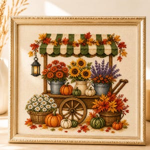May include: A framed cross-stitch embroidery depicting a wooden cart under a green and white striped awning. The cart holds buckets of sunflowers, orange flowers, and lavender, surrounded by pumpkins, white daisies, and autumn foliage.