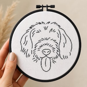 May include: A circular black embroidery hoop containing white Aida fabric with a black cross-stitch outline of a shaggy dog face. The dog design features floppy ears, a textured coat, a rounded nose, and a tongue hanging out of its mouth.