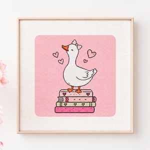 May include: A framed cross-stitch embroidery featuring a white duck with an orange beak and feet, wearing a small bow on its head. The duck stands on a stack of four books against a pink background decorated with several small floating hearts.