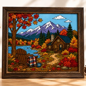 May include: A framed cross-stitch embroidery depicting a mountain landscape in autumn. The scene features a wooden cabin, a lake, a large tree with orange leaves, and an Adirondack chair with a plaid blanket and a blue mug, all in a dark wood frame.
