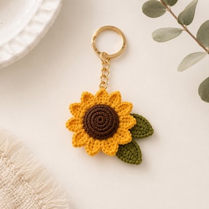 May include: A crocheted sunflower keychain with a gold-colored keyring and chain. The sunflower has yellow petals, a brown center, and two green leaves. The keychain is on a white surface with a decorative plate and greenery in the background.