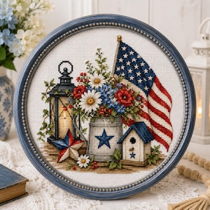 May include: A circular cross-stitch embroidery in a blue frame featuring a black lantern, a metal bucket with a blue star, a birdhouse, a bouquet of red, white, and blue flowers, and an American flag, all set against a white fabric background.