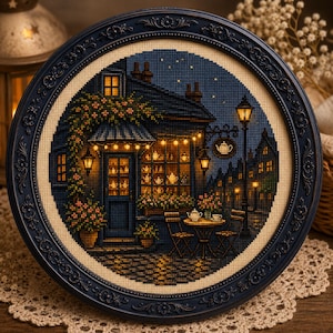 May include: A circular cross-stitch embroidery depicting a cozy tea shop at night, framed in an ornate dark blue border. The scene features a storefront with glowing windows, string lights, a street lamp, and a small table with tea service.