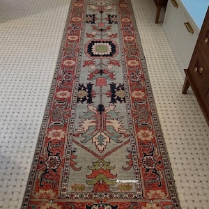 May include: A long, rectangular runner rug featuring a light blue central field with a symmetrical pattern of floral and geometric motifs in shades of red, navy blue, green, and cream, enclosed by a wide, ornate red border with intricate detailing.