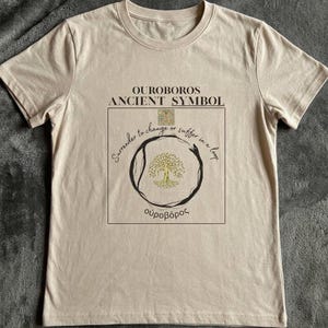 Op de afbeelding: Beige t-shirt met de tekst "OUROBOROS ANCIENT SYMBOL" boven een cirkelvormig ontwerp met een boom en het Griekse woord "ουροβόρος". Het ontwerp bevat de zin "Surrender to change or suffer in a loop."