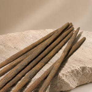 May include: Close-up of several brown incense sticks with a textured, speckled appearance. The incense sticks are arranged on a light-colored, rough stone surface, with a neutral background.