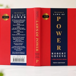 May include: A hardcover book titled The Concise 48 Laws of Power by Robert Greene and Joost Elffers. The cover features a bold red and dark blue design with gold lettering. The back includes a gold SND seal and a barcode with the number 2217 A.