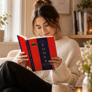 May include: A hardcover book titled The Concise 48 Laws of Power by Robert Greene with a bright red cover, a dark blue vertical center stripe, and gold lettering. The book is held open, displaying the front cover design and the spine text clearly.