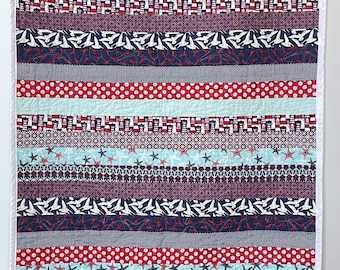 May include: A red, white, and blue striped baby quilt with a nautical theme. The quilt features anchors, stars, and sailboats.