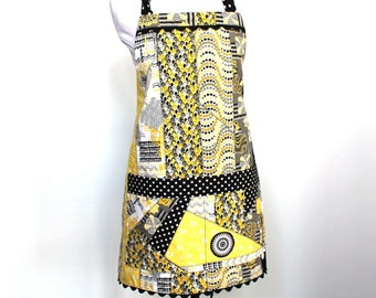 May include: A yellow and black patchwork apron with a black polka dot trim and a black scalloped edge. The apron has a black and white polka dot pocket.