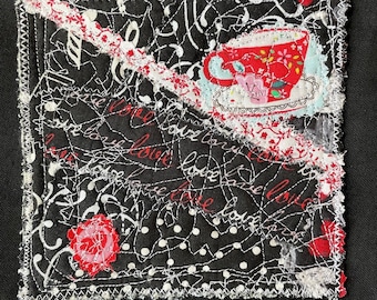 May include: A black fabric square with white stitching and a red and white teacup design. The fabric is decorated with the words "love" and "be love" in a cursive font.
