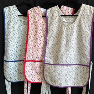 May include: Three floral print aprons with blue, red, and purple trim hang against a black background. Each apron has a pocket and ties. The aprons feature a cream-colored background with a small blue or red floral pattern.