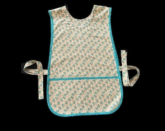 May include: A beige apron with a teal floral pattern and teal trim. The apron has a pocket and ties at the waist. The apron is sleeveless and has a rounded neckline. The apron is made of fabric.