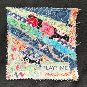 May include: A small, square fabric art piece with a patchwork design. The piece features various colorful fabric scraps stitched together in diagonal stripes. The word "PLAYTIME" is printed on a white strip of fabric. The edges are frayed, giving it a handmade look.