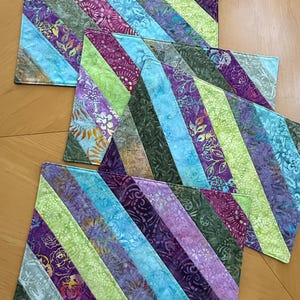 May include: A set of three rectangular placemats featuring a diagonal striped pattern. The fabric displays a variety of batik-style prints in shades of purple, lime green, teal, and dark olive, with floral and abstract motifs throughout the design.