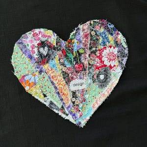 May include: A heart-shaped fabric patch constructed from various colorful textile scraps featuring floral, cherry, and geometric patterns. The piece is held together with white topstitching and includes a small white label with the word design.