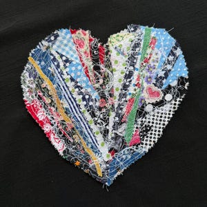May include: A handmade, fabric heart-shaped item with a patchwork design. The heart is composed of various fabric strips in a range of colors and patterns, including floral, polka dots, and stripes. The background is solid black.