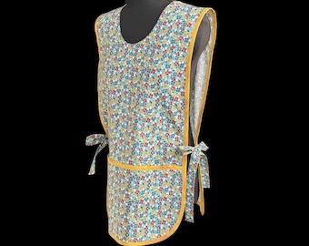 May include: A floral print apron with a yellow trim and ties. The apron features a pattern of small blue, red, and yellow flowers on a light blue background. The apron is displayed on a black mannequin.