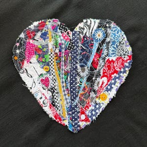 May include: A heart-shaped fabric patch composed of colorful, patterned fabric strips. The strips are sewn together with visible stitching, creating a patchwork design. The background is a solid black fabric.