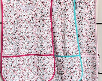 May include: Three sleeveless aprons with a floral print and contrasting trim. The aprons have a large pocket on the front and tie straps.