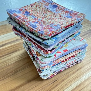 May include: A stack of colorful, square fabric coasters. The coasters feature various floral and patterned designs in shades of blue, pink, red, and yellow. They are stacked on a wooden surface.