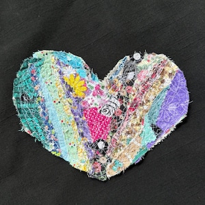 May include: A heart-shaped fabric applique constructed from various strips of multicolored patterned textiles. The design features floral, polka dot, and geometric prints in shades of blue, yellow, pink, purple, and green, held together by white stitching.