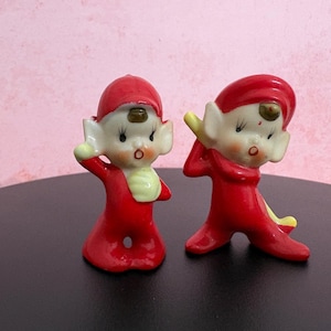 Op de afbeelding: Twee keramische elfenbeeldjes in felrode outfits met bijpassende rode mutsen. Elke elf heeft een bleek gezicht, grote puntige oren en geschilderde gelaatstrekken. De ene elf staat rechtop met een hand omhoog, terwijl de andere licht opzij leunt.