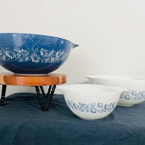 May include: A set of three vintage Pyrex mixing bowls. The largest bowl is a deep blue with a white floral design, resting on a wooden stand with black metal legs. Two smaller white bowls with blue floral patterns are also visible.