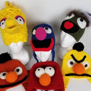 May include: A collection of six handmade character hats, including Big Bird, Elmo, and Oscar the Grouch. The hats are crocheted in bright colors like yellow, red, and blue, with detailed facial features and pom-poms.