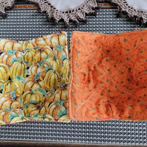 May include: Two fabric bowl cozies. One is orange with a pattern of acorns and leaves. The other is yellow with a pattern of pumpkins and leaves.