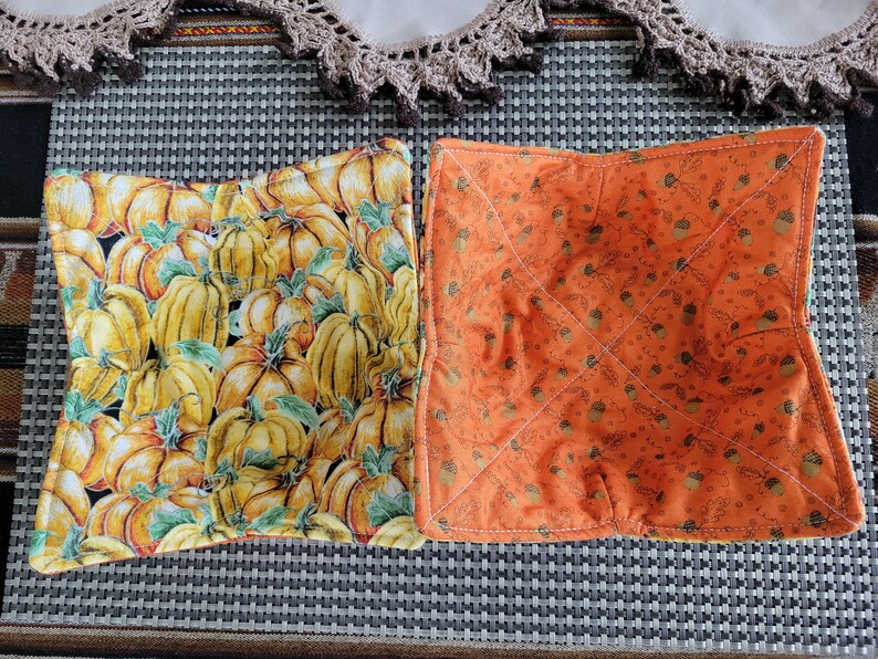 May include: Two fabric bowl cozies. One is orange with a pattern of acorns and leaves. The other is yellow with a pattern of pumpkins and leaves.