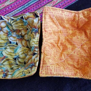 May include: Two square fabric pot holders. One is yellow with a red pattern and the other is yellow with a banana and pumpkin print.