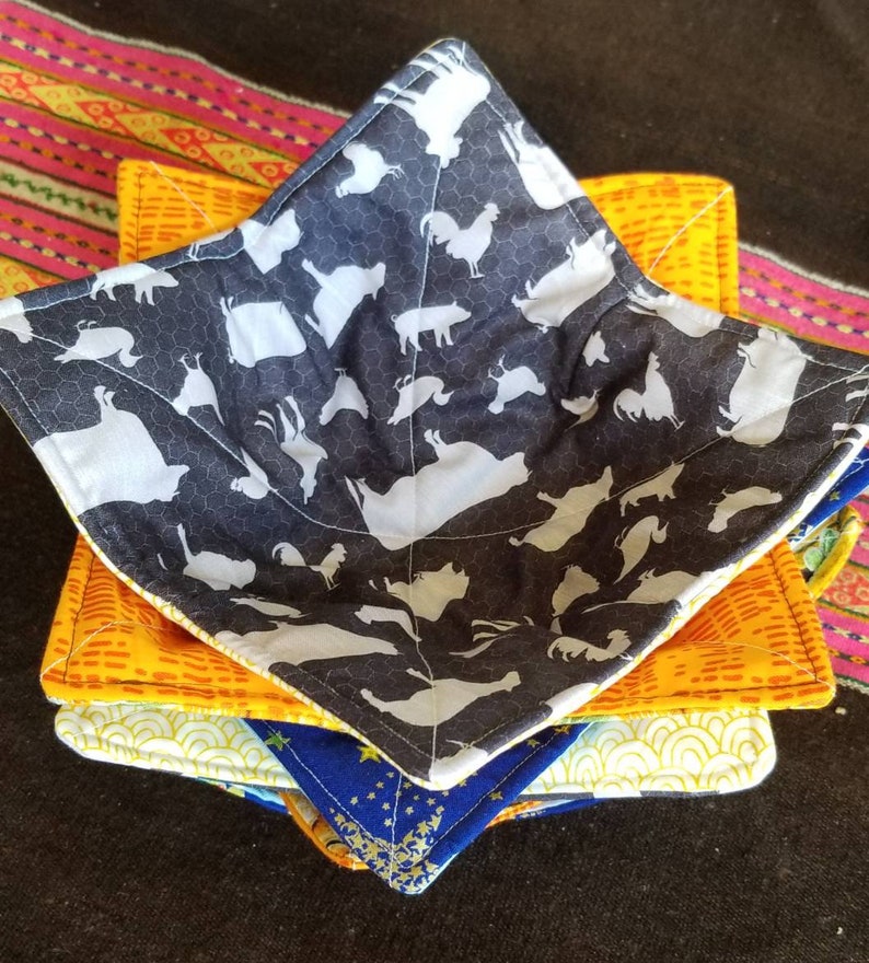 May include: A set of four quilted bowl cozies in various patterns. The top cozy features a black and white pattern of farm animals, the second cozy is orange with a yellow geometric pattern, the third cozy is yellow with a white geometric pattern, and the bottom cozy is blue with a gold star pattern.