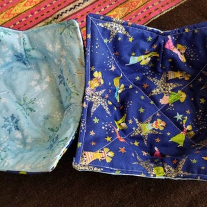 May include: Two blue fabric bowl cozies. One is a light blue with a snowflake pattern. The other is a dark blue with a gold star and glitter pattern with a whimsical illustration of people.