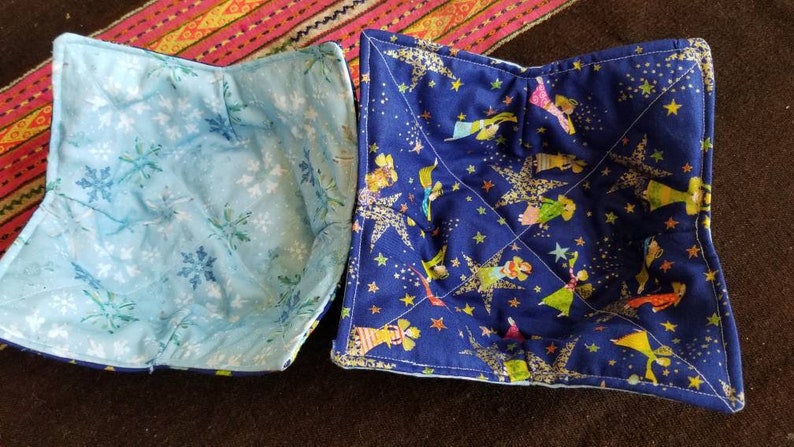 May include: Two blue fabric bowl cozies. One is a light blue with a snowflake pattern. The other is a dark blue with a gold star and glitter pattern with a whimsical illustration of people.