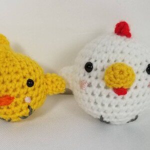 May include: Two crocheted chicken plushies. One is yellow with a red beak and the other is white with a yellow beak and a red comb.